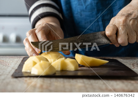 A senior person uses a vintage steel knife to slice peeled potatoes on a wooden board, emphasizing traditional food preparation and home cooking methods in a cozy domestic kitchen A senior person uses a vintage steel knife to slice peeled potatoes on a wooden board, emphasizing traditional food preparation and home cooking methods in a cozy domestic kitchen 140676342