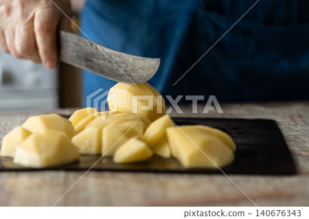 A hand uses a sharp knife to cut raw potatoes on a dark wooden board, conveying the preparation of fresh food for a healthy meal in a cozy home kitchen setting for daily cooking 140676343