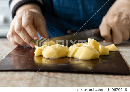 A pair of hands slices raw yellow potatoes on a wooden board using a sharp steel knife, emphasizing home cooking preparation, healthy nutrition, and culinary skill in a kitchen setting A pair of hands slices raw yellow potatoes on a wooden board using a sharp steel knife, emphasizing home cooking preparation, healthy nutrition, and culinary skill in a kitchen setting 140676348
