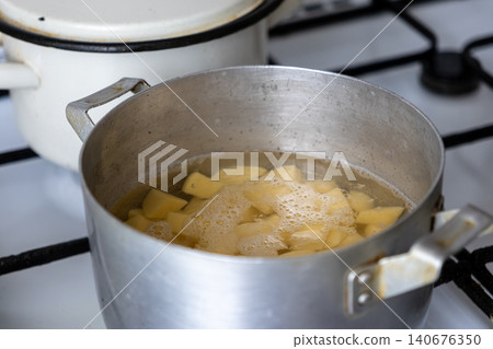 Chopped raw potato chunks boil in a metal pot on a white stove, representing the home culinary process of preparing nutritious food for a simple meal within a modern kitchen setting Chopped raw potato chunks boil in a metal pot on a white stove, representing the home culinary process of preparing nutritious food for a simple meal within a modern kitchen setting 140676350