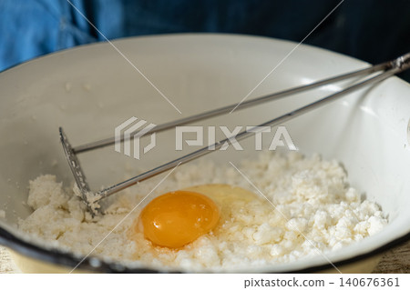 Fresh cottage cheese with a raw egg yolk in a white bowl being mashed with a metal tool for a homemade dairy baking preparation in a bright kitchen setting for healthy cooking recipes Fresh cottage cheese with a raw egg yolk in a white bowl being mashed with a metal tool for a homemade dairy baking preparation in a bright kitchen setting for healthy cooking recipes 140676361