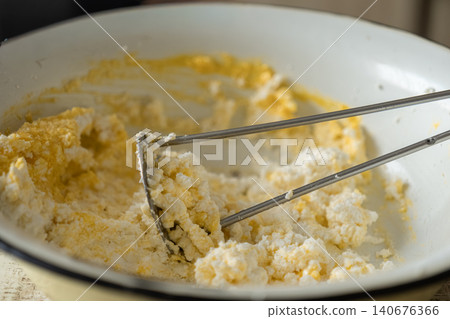A manual potato masher crushing cottage cheese with egg yolk in a rustic white bowl, capturing a home cooking preparation process for delicious healthy protein snacks and light meals 140676366