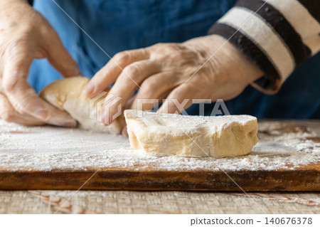 An elderly person shapes soft raw dough on a wooden board covered in white flour, representing traditional artisanal baking techniques for a cozy homemade meal in the rustic kitchen An elderly person shapes soft raw dough on a wooden board covered in white flour, representing traditional artisanal baking techniques for a cozy homemade meal in the rustic kitchen 140676378