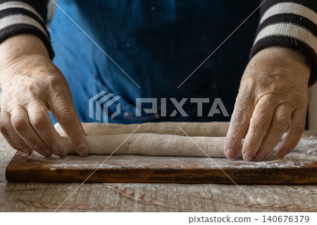 A pair of wrinkled hands kneads dough on a wooden board, emphasizing the rustic charm of handmade baking and traditional artisanal culinary practices within a cozy kitchen environment A pair of wrinkled hands kneads dough on a wooden board, emphasizing the rustic charm of handmade baking and traditional artisanal culinary practices within a cozy kitchen environment 140676379