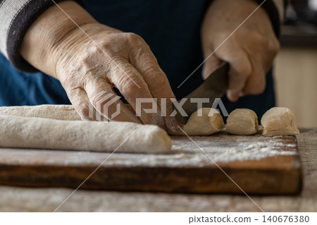 An elderly person cuts pieces of dough on a wooden board with a sharp kitchen knife to prepare traditional food, emphasizing handcrafted culinary arts in a rustic domestic setting An elderly person cuts pieces of dough on a wooden board with a sharp kitchen knife to prepare traditional food, emphasizing handcrafted culinary arts in a rustic domestic setting 140676380