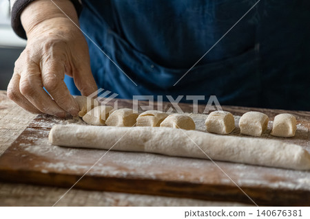 A pair of senior hands cuts pieces of raw dough on a wooden board to create handmade gnocchi, emphasizing authentic culinary traditions and a nurturing home cooking kitchen atmosphere A pair of senior hands cuts pieces of raw dough on a wooden board to create handmade gnocchi, emphasizing authentic culinary traditions and a nurturing home cooking kitchen atmosphere 140676381