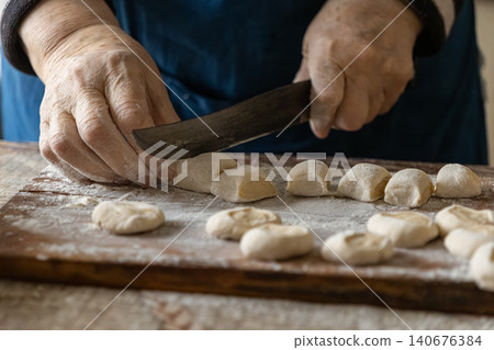A senior person cuts fresh dough into small pieces for handmade dumplings on a floured wooden board, capturing the traditional craft of home cooking and authentic culinary heritage A senior person cuts fresh dough into small pieces for handmade dumplings on a floured wooden board, capturing the traditional craft of home cooking and authentic culinary heritage 140676384