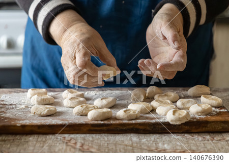 A senior person carefully shaping homemade dough into small round dumplings on a floured wooden board, representing traditional cooking methods and the warmth of a rustic kitchen home 140676390