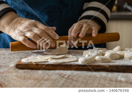 A senior person rolls fresh dough with a wooden pin on a floury board to prepare homemade dumplings, capturing traditional culinary craft and authentic kitchen skills in close detail 140676398