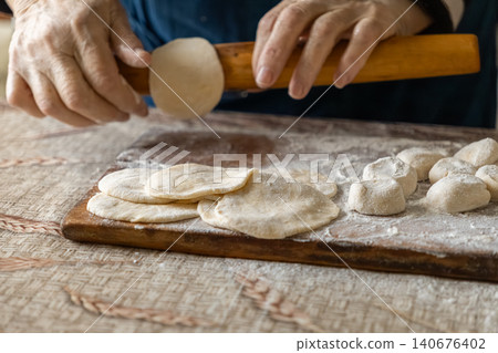 Senior person rolls fresh dough circles with a wooden pin on a floured board to prepare homemade dumplings, capturing the traditional craft of cooking healthy and authentic food Senior person rolls fresh dough circles with a wooden pin on a floured board to prepare homemade dumplings, capturing the traditional craft of cooking healthy and authentic food 140676402