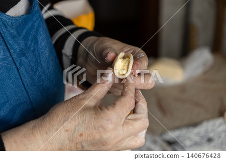 A senior woman folds dough to prepare handmade cheese dumplings, reflecting traditional culinary skills and the authentic passion of homemade cooking in a cozy kitchen environment A senior woman folds dough to prepare handmade cheese dumplings, reflecting traditional culinary skills and the authentic passion of homemade cooking in a cozy kitchen environment 140676428
