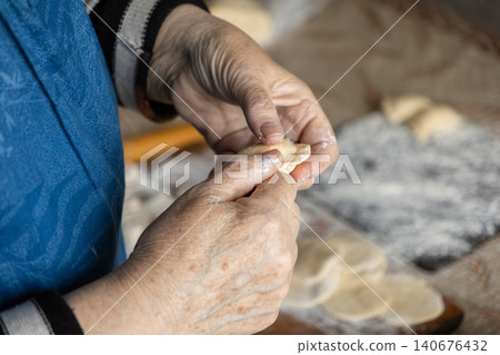 A senior person preparing handmade dumplings by pinching dough edges carefully at a rustic kitchen table, illustrating a warm culinary tradition focused on fresh authentic home cooking A senior person preparing handmade dumplings by pinching dough edges carefully at a rustic kitchen table, illustrating a warm culinary tradition focused on fresh authentic home cooking 140676432