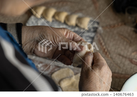 An elderly person shaping handmade dumplings by folding dough at a kitchen table, capturing traditional culinary skills and the warmth of preparing a homemade meal for the family 140676463