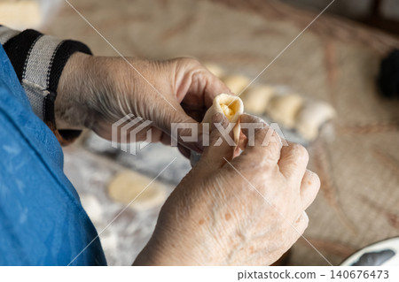 Elderly hands meticulously folding dough to prepare homemade dumplings at a kitchen table, capturing the traditional craft of artisan food cooking in a cozy and warm domestic setting Elderly hands meticulously folding dough to prepare homemade dumplings at a kitchen table, capturing the traditional craft of artisan food cooking in a cozy and warm domestic setting 140676473