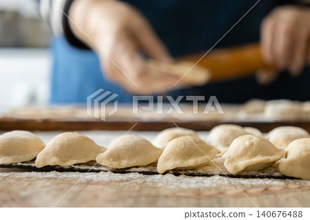 Uncooked dumplings sit on a floured wooden surface while a person prepares more pastry dough in the background, creating an authentic scene of traditional homemade meal preparation Uncooked dumplings sit on a floured wooden surface while a person prepares more pastry dough in the background, creating an authentic scene of traditional homemade meal preparation 140676488