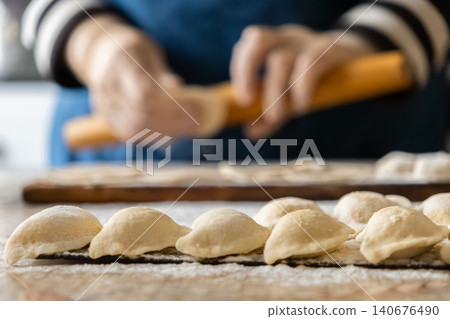 A chef prepares traditional dumplings by rolling dough on a floured wooden surface, capturing the authentic essence of handmade craft in a warm and cozy home kitchen environment A chef prepares traditional dumplings by rolling dough on a floured wooden surface, capturing the authentic essence of handmade craft in a warm and cozy home kitchen environment 140676490