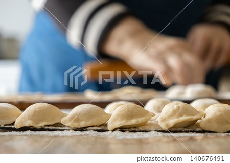 Fresh handmade dumplings lined up on a floured wooden surface while a person prepares dough in the background to convey a warm culinary tradition of home cooking and artisan food Fresh handmade dumplings lined up on a floured wooden surface while a person prepares dough in the background to convey a warm culinary tradition of home cooking and artisan food 140676491
