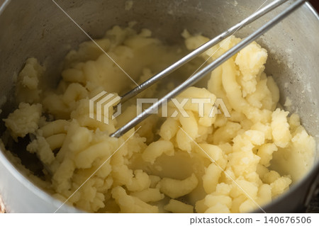 Boiled potato being mashed inside a metallic pot with a steel tool to prepare a filling for handmade savory dumplings, showcasing the rustic cooking process in a home kitchen Boiled potato being mashed inside a metallic pot with a steel tool to prepare a filling for handmade savory dumplings, showcasing the rustic cooking process in a home kitchen 140676506
