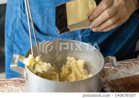 A chef adding spice to mashed potato filling for dumplings in a rustic metal pot, captured during the careful preparation of authentic homemade savory dough dishes in a kitchen A chef adding spice to mashed potato filling for dumplings in a rustic metal pot, captured during the careful preparation of authentic homemade savory dough dishes in a kitchen 140676507