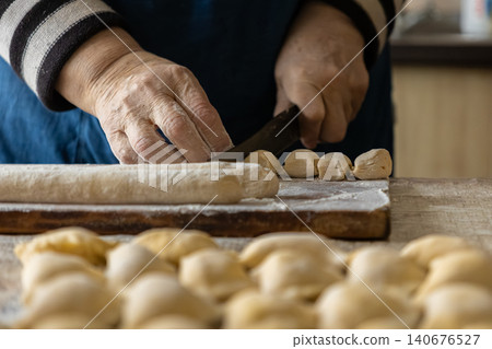 A senior person slices a dough cylinder with a sharp knife on a wooden board, preparing fresh handmade dumplings in a home kitchen setting that evokes warmth and culinary tradition A senior person slices a dough cylinder with a sharp knife on a wooden board, preparing fresh handmade dumplings in a home kitchen setting that evokes warmth and culinary tradition 140676527