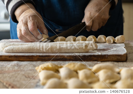 A person cuts raw dough into small pieces for handmade dumplings on a floury wooden board, demonstrating a traditional culinary craft in a warm home kitchen for authentic meal prep A person cuts raw dough into small pieces for handmade dumplings on a floury wooden board, demonstrating a traditional culinary craft in a warm home kitchen for authentic meal prep 140676528