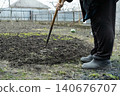 A senior person uses a metal hoe to cultivate dark fertile soil in a rural backyard garden plot, embodying a dedicated lifestyle of spring planting, seasonal farming and earthy growth 140676707