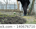 A senior person stands in a vegetable garden holding a long handled metal hoe during spring, conveying the quiet persistence of agricultural labor and seasonal preparation of soil 140676710