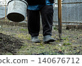 A gardener walking with a plastic bucket and a metal hoe in a backyard, embodying agricultural effort, outdoor maintenance, and sustainable lifestyle practices in a rural setting 140676712