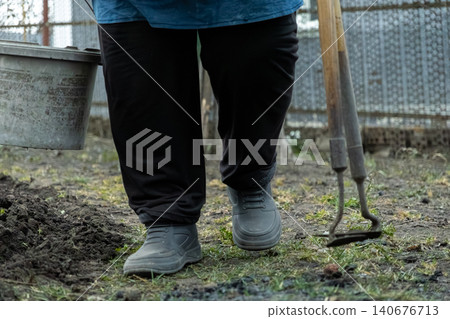 A gardener walking across a dirt backyard plot while carrying a plastic bucket and a hand hoe, demonstrating active garden maintenance during the spring season for a healthy lawn A gardener walking across a dirt backyard plot while carrying a plastic bucket and a hand hoe, demonstrating active garden maintenance during the spring season for a healthy lawn 140676713