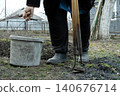 A senior person holding a worn bucket and metal garden hoe in a backyard soil patch, representing the physical labor of spring planting and seasonal maintenance in a rural landscape 140676714