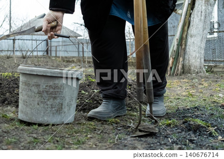 A senior person holding a worn bucket and metal garden hoe in a backyard soil patch, representing the physical labor of spring planting and seasonal maintenance in a rural landscape A senior person holding a worn bucket and metal garden hoe in a backyard soil patch, representing the physical labor of spring planting and seasonal maintenance in a rural landscape 140676714