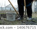 A gardener in rubber boots uses a metal hoe beside a plastic bucket in a vegetable plot, demonstrating manual soil preparation techniques for seasonal spring planting and cultivation 140676715