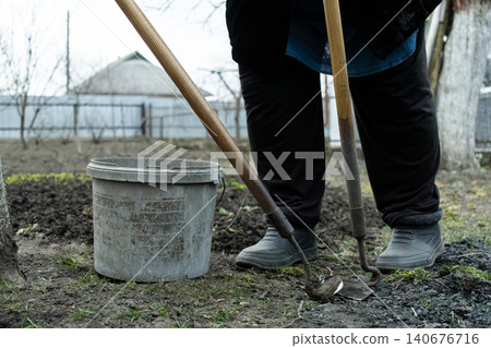 A gardener uses a sharp hoe to cultivate rich soil near a plastic bucket in an outdoor garden plot, depicting rural spring agricultural labor and preparation for planting crops A gardener uses a sharp hoe to cultivate rich soil near a plastic bucket in an outdoor garden plot, depicting rural spring agricultural labor and preparation for planting crops 140676716