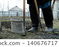 A gardener holds two hoes beside a grey plastic bucket in a vegetable patch, demonstrating the essential labor of spring soil preparation for a productive rural home garden harvest 140676717