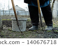 A gardener stands in a vegetable patch with steel hoes and a plastic bucket, preparing soil for spring planting while demonstrating traditional farming and rural lifestyle tasks 140676718