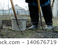 A gardener uses a pair of metal hand hoes near a plastic bucket on fresh soil to cultivate a rural vegetable plot, reflecting humble agricultural work and seasonal land care 140676719