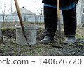 A gardener standing with a plastic bucket and a manual hoe in a muddy vegetable plot during early spring, emphasizing traditional agricultural preparation for planting seasonal crops 140676720