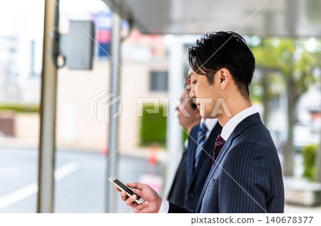 A man in a suit is using his smartphone while waiting for a bus at a bus stop. A man in a suit is using his smartphone while waiting for a bus at a bus stop. 140678377