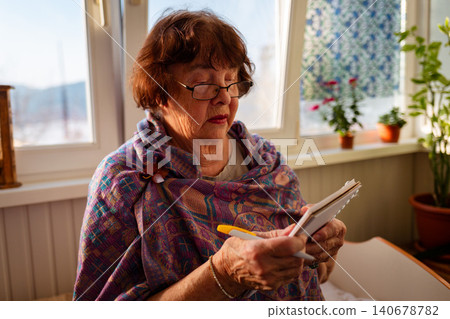 Elderly Caucasian woman documenting memories and experiences in a notebook near a sunny window. Reflection and lifelong learning concept. Mid shot Elderly Caucasian woman documenting memories and experiences in a notebook near a sunny window. Reflection and lifelong learning concept. Mid shot 140678782