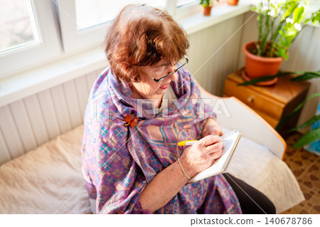 Caucasian grandmother taking educational notes while enjoying daylight indoors. Learning and creativity concept. Healthy aging. Top view Caucasian grandmother taking educational notes while enjoying daylight indoors. Learning and creativity concept. Healthy aging. Top view 140678786
