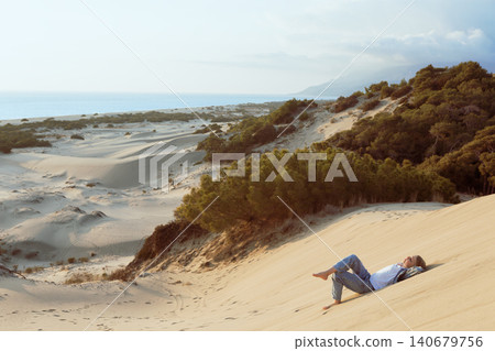Young girl relaxes on a steep sand dune at Patara Beach, Turkey. Minimal coastal landscape, warm sunlight and calm travel mood during summer vacation by the sea. Young girl relaxes on a steep sand dune at Patara Beach, Turkey. Minimal coastal landscape, warm sunlight and calm travel mood during summer vacation by the sea. 140679756
