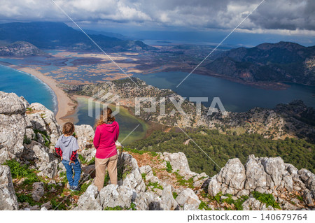 Young mother and son stand on mountain viewpoint above Dalyan, Turkey, overlooking river delta, lagoon, Iztuzu Turtle beach. Scenic travel moment highlighting family adventure and nature exploration. Young mother and son stand on mountain viewpoint above Dalyan, Turkey, overlooking river delta, lagoon, Iztuzu Turtle beach. Scenic travel moment highlighting family adventure and nature exploration. 140679764