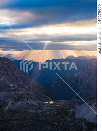 Powerful sunbeams break through dramatic storm clouds above the Tre Cime di Lavaredo region in the Dolomites, Italy. Aerial view of rugged alpine terrain illuminated by golden evening light. 140679909