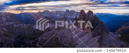 Aerial view of the iconic Tre Cime di Lavaredo rock towers at dusk in the Dolomites, Italy. Rugged limestone peaks rise above alpine valleys beneath a pastel evening sky. Aerial view of the iconic Tre Cime di Lavaredo rock towers at dusk in the Dolomites, Italy. Rugged limestone peaks rise above alpine valleys beneath a pastel evening sky. 140679910