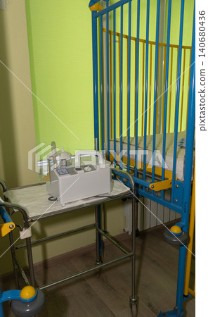 Medical monitoring device on a stainless steel trolley next to a colorful baby crib in a clinic. Medical monitoring device on a stainless steel trolley next to a colorful baby crib in a clinic. 140680436