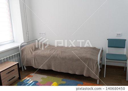 Minimalist single metal bed with tan blanket in bright white room with chair and colorful play mat Minimalist single metal bed with tan blanket in bright white room with chair and colorful play mat 140680450