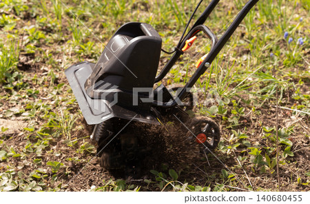 Electric garden cultivator tilling soil in green backyard during spring season for planting vegetables. Electric garden cultivator tilling soil in green backyard during spring season for planting vegetables. 140680455