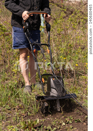 Man operating an electric garden tiller to cultivate soil in a backyard garden during spring. 140680465