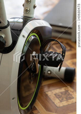 Close up of stationary exercise bike pedal with black strap on wooden floor in bright room. Close up of stationary exercise bike pedal with black strap on wooden floor in bright room. 140680478