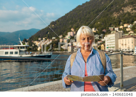 Mature woman traveler searching for direction with city map against mountain lake background Mature woman traveler searching for direction with city map against mountain lake background 140681048